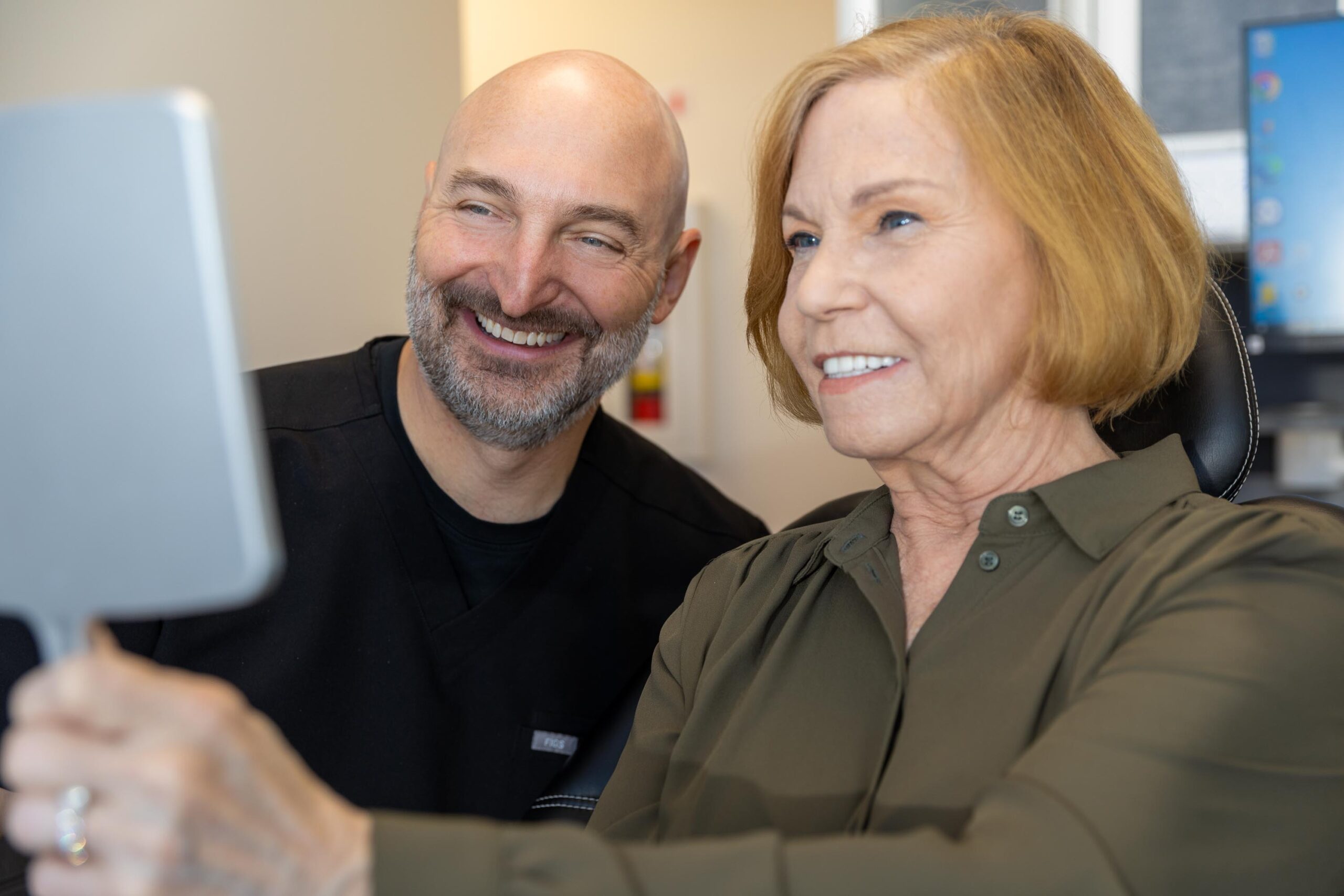 Dr. Fetsch and his patient smiling at her reflection in a hand held mirror after her full mouth dental implant procedure.