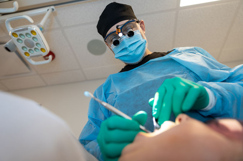 A dentist in surgical scrubs performs a dental implant procedure on a sedated patient in a modern, well-equipped dental office.